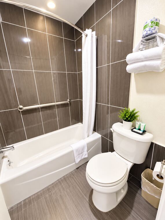 Bathroom with a white tub and shower against brown tile, folded towels on a chrome shelf and toiletries on the toilet tank.