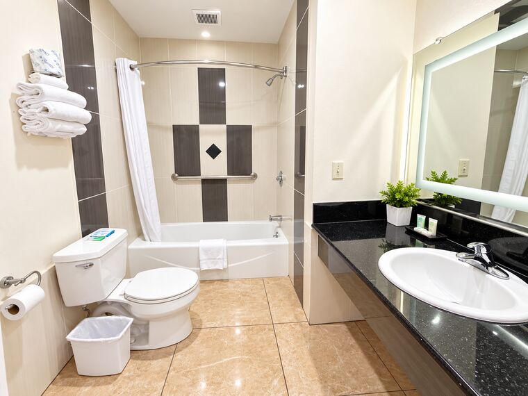Bathroom with a tub and shower in cream and charcoal tile, a lighted mirror over a black granite vanity and stacked white towels.