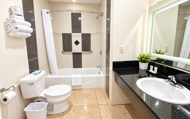 A guest bathroom with a lighted mirror over a black granite vanity and a tub and shower in cream and charcoal tile.